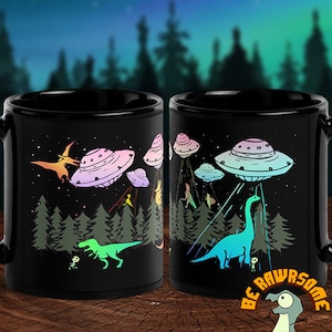 May include: Two black coffee mugs with a colorful graphic of dinosaurs and UFOs in a forest setting. The text "Be Rawsome" is on the bottom of the mug.