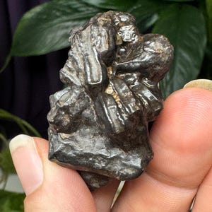 Puede incluir: Una piedra oscura, de forma irregular, de aspecto metálico, con una superficie rugosa y texturizada. La piedra tiene una mezcla de tonos grises oscuros y marrones, con algunas áreas brillantes. La forma general es orgánica y algo abstracta, posiblemente un espécimen mineral.