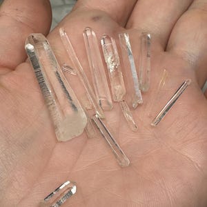 May include: A collection of clear quartz crystal points of varying sizes and shapes. The crystals are held in an open palm, showcasing their translucent quality and natural formations. The crystals are a light, neutral color.
