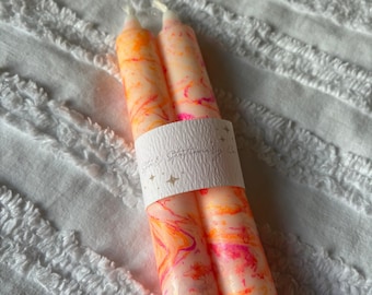 May include: Two taper candles featuring a marbled pattern of bright orange, hot pink, and white wax. The candles are bound together by a white paper band printed with the text Star Stationery Co. and small star icons, with visible white wicks.