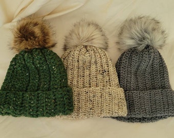 May include: Three knitted winter hats with faux fur pom poms. The hats are green, beige, and gray.