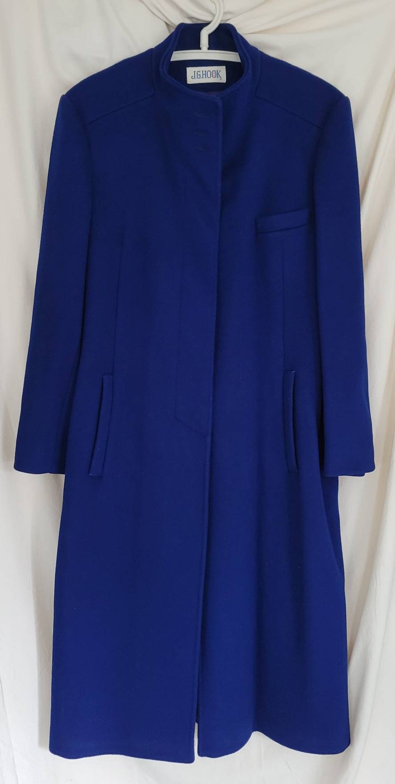 May include: A long, royal blue coat with a mandarin collar and a single button fastening. The coat has a small pocket on the left side and two side pockets. The label on the coat reads "JG HOOK."