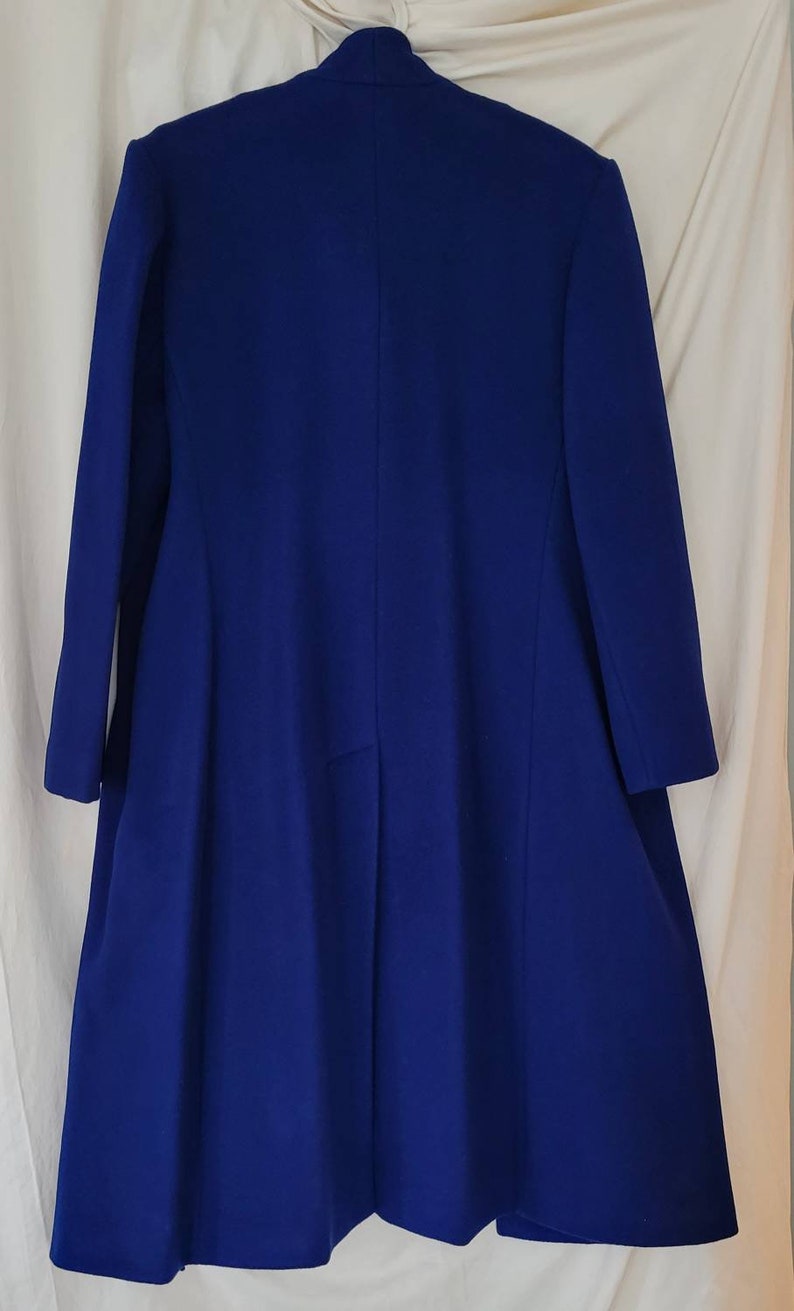 May include: A long, royal blue coat with a simple, elegant design. The coat has a high collar and long sleeves. The fabric is smooth and appears to be wool.