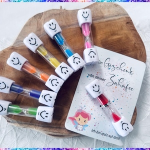 Könnte beinhalten: Ein Set aus acht bunten, zahnförmigen Sanduhren mit einem Smiley auf jeder. Die Sanduhren sind in einem Halbkreis auf einer Holzoberfläche angeordnet. Eine Karte mit dem Text "Ein Geschenk von deiner Zahnfee" und "Ich bin stolz auf dich!" befindet sich im Hintergrund.