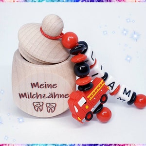 Könnte beinhalten: Eine Holzkiste mit dem Text "Meine Milchzähne" und zwei Zahnsymbolen. Die Kiste ist offen und eine rot-schwarze Perlenkette mit einem roten Feuerwehrauto-Anhänger hängt aus der Kiste. Die Kette hat weiße Buchstabenperlen, die "AM" buchstabieren.