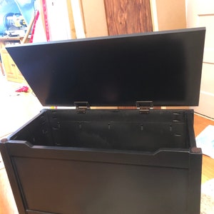 May include: Black wooden storage chest with a hinged lid. The lid is open, revealing the empty interior of the chest.