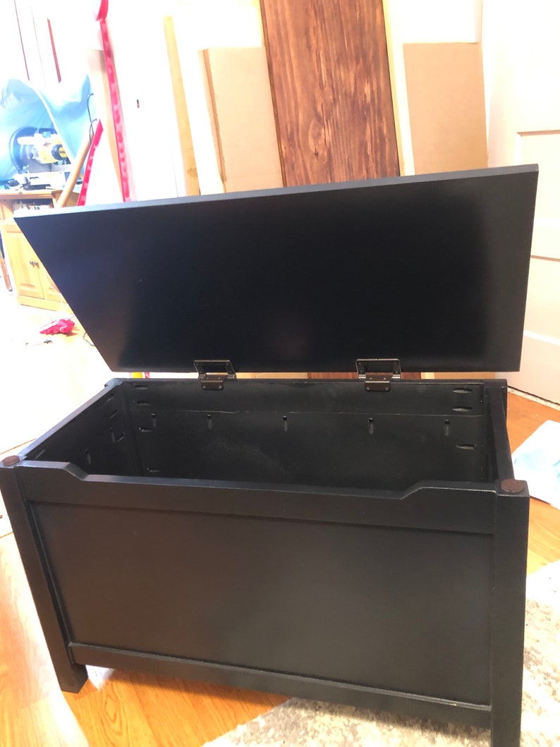 May include: Black wooden storage chest with a hinged lid. The lid is open, revealing the empty interior of the chest.