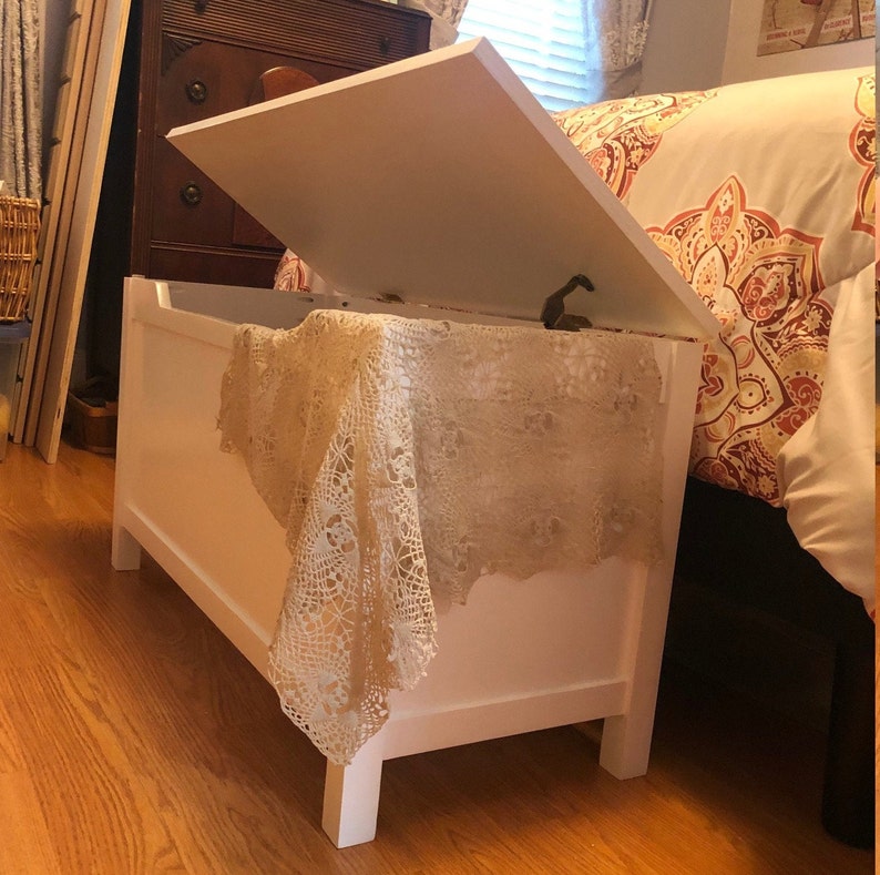 May include: White wooden toy chest with a hinged lid that is open. The chest is sitting on a wooden floor and has a crocheted tablecloth draped over it.