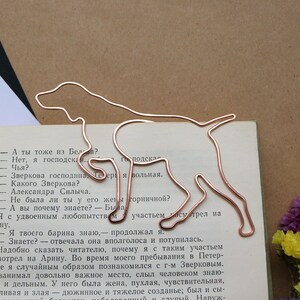 Op de afbeelding: Een bladwijzer van koperdraad in de vorm van een hond, met een boekpagina op de achtergrond. De bladwijzer is een silhouet van een hond met zijn kop naar links gedraaid en zijn staart opgerold. De boekpagina bevat tekst in het Russisch.