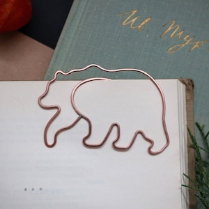 May include: A copper-colored wire bear-shaped bookmark rests on the pages of an open book. The bookmark is a simple outline of a bear. The book has text in a foreign language. The background includes a vintage book and greenery.