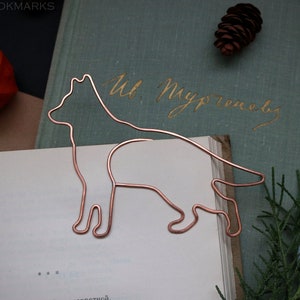 May include: A copper wire bookmark shaped like a dog standing on its four legs. The bookmark is on top of a book with the text "Ut Myprousley" written in gold ink on the cover.
