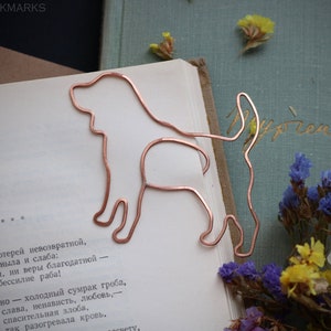 May include: A copper wire bookmark shaped like a dog, sitting on a book with text in Russian. The bookmark is a simple silhouette of a dog with a long tail.