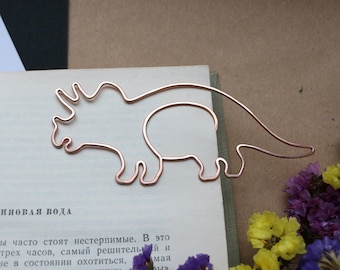 May include: A rose gold metal bookmark shaped like a dinosaur. The bookmark is on top of a book with text.