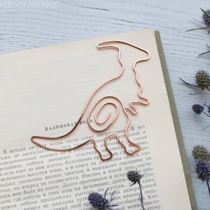 Puede incluir: Un marcapáginas de alambre de cobre con forma de dinosaurio. El marcapáginas está sobre un libro con texto en ruso. El libro está sobre una superficie de madera blanca con flores secas.
