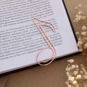May include: A rose gold metal bookmark shaped like a musical note. The bookmark is lying open on a book with white flowers in the background.