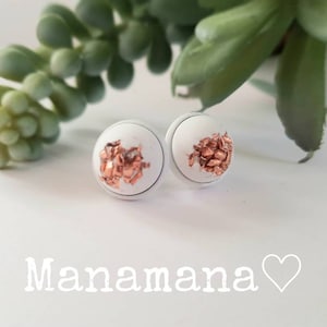 Könnte beinhalten: Ein Paar weiße Ohrstecker mit kupferfarbenen Akzenten. Die Ohrringe haben eine runde Form und sind vor einem verschwommenen Hintergrund mit einer Sukkulentenpflanze platziert. Das Wort "Manamana" ist in weißer Schrift geschrieben.