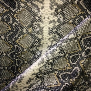 May include: Close-up of a snakeskin pattern fabric. The fabric features a repeating pattern of scales in shades of black, grey, white, and gold. The texture appears smooth and slightly reflective, with a diagonal fold visible.
