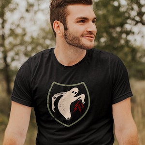 May include: A black t-shirt with a green and white ghost graphic. The ghost is holding a red leaf. The shirt is being worn by a person sitting on a wooden fence.