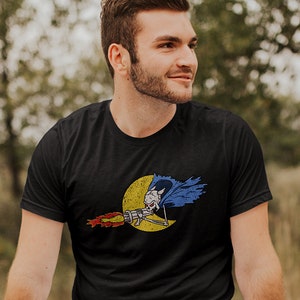 May include: A black t-shirt with a graphic of a skeleton riding a rocket with a blue cape and yellow moon in the background.