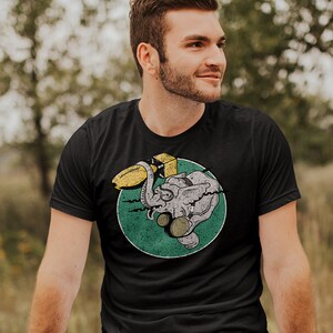 May include: A black t-shirt with a distressed green circle graphic of a cartoon elephant holding a yellow banana. The elephant is wearing a yellow helmet.