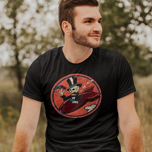 May include: Black t-shirt featuring a distressed circular graphic of a skeleton in a top hat riding a red rocket, holding a gun. The design has a vintage, worn look. The shirt is paired with blue jeans.
