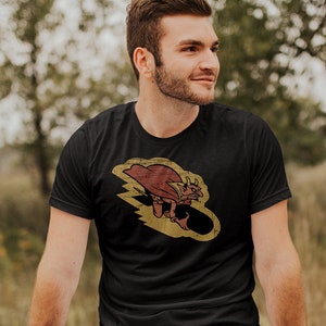 May include: A black t-shirt with a vintage-style graphic of a cartoon devil riding a bomb. The graphic is yellow, red, and brown.