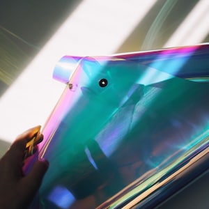 Puede incluir: Una bolsa de plástico transparente holográfica iridiscente con una cremallera dorada. La bolsa está doblada por la mitad y sostenida por una mano.