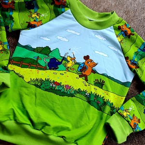 Könnte beinhalten: Grünes Langarm-Sweatshirt mit einer Cartoon-Szene mit einer Maus, einer Ente und einem Elefanten auf einem Weg. Die Ärmel und der Saum sind einfarbig grün, der Körper hat einen hellblauen Himmel. Der Text "nice handcrafted" befindet sich oben rechts.