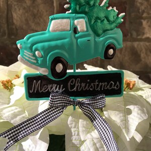 May include: A turquoise truck carrying a green Christmas tree with white snow on the branches. The truck has a black chalkboard sign that says "Merry Christmas" and a black and white gingham ribbon tied around the sign.