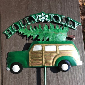 May include: A green metal Christmas decoration featuring a vintage station wagon with a Christmas tree on top. The car has wood paneling and the words "HOLLY JOLLY" are above the car.