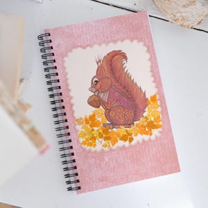 Könnte beinhalten: Ein spiralgebundenes Notizbuch mit rosa Einband, das eine detaillierte Illustration eines Eichhörnchens mit einer Nuss zeigt, umgeben von Herbstblättern. Das Eichhörnchen ist in Rot- und Brauntönen gehalten, mit einem weißen, gezackten Rand.