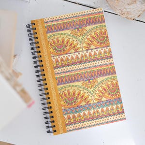 May include: Spiral-bound notebook with a colourful cover. The cover has a repeating pattern of eye-like shapes, triangles, and geometric designs in yellow, orange, pink, and blue. The notebook's spine is mustard yellow.