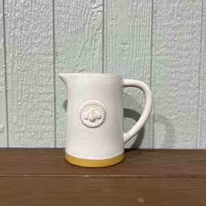 May include: A white ceramic pitcher with a yellow base and a handle. The pitcher features a raised bee emblem on the front. The background is a light blue wooden wall. The pitcher is a decorative kitchen item.