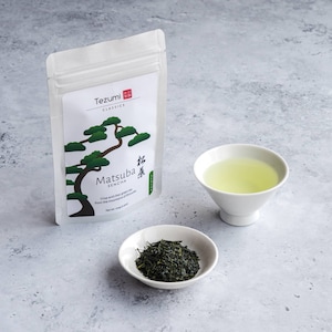 Könnte beinhalten: Ein weißer Beutel mit Tezumi Classics Matsuba Sencha Tee mit einer Baumillustration, eine kleine weiße Schale mit losem Tee und eine weiße Teetasse gefüllt mit grünem Tee. Der Text auf dem Beutel lautet "Knackiger und klarer grüner Tee aus den Bergen von Shizuoka."