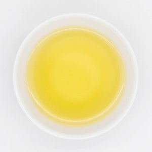 Puede incluir: Vista superior de un cuenco de cer&aacute;mica blanca lleno de un l&iacute;quido amarillo dorado. El cuenco tiene una forma simple y redondeada y se encuentra sobre un fondo blanco liso. El l&iacute;quido parece suave y transparente.