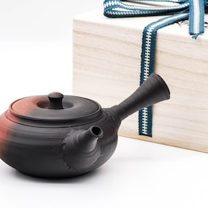 Può includere: Una teiera in ceramica nera e rossa con un beccuccio e un coperchio. La teiera è appoggiata su una superficie bianca accanto a una scatola di legno con un nastro blu.