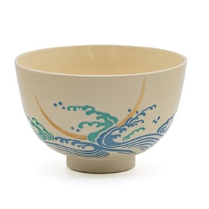 May include: Cream-colored ceramic bowl with a footed base. The bowl features a wave design in shades of blue and teal, accented with gold details. The interior is a lighter cream color.