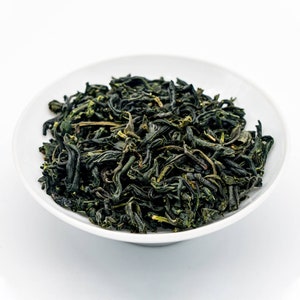 Puede incluir: Un cuenco de cer&aacute;mica blanco lleno de hojas de t&eacute; verde secas. Las hojas de t&eacute; son de color verde oscuro con algunos reflejos verde amarillentos m&aacute;s claros. El cuenco es redondo y est&aacute; sobre un fondo blanco.