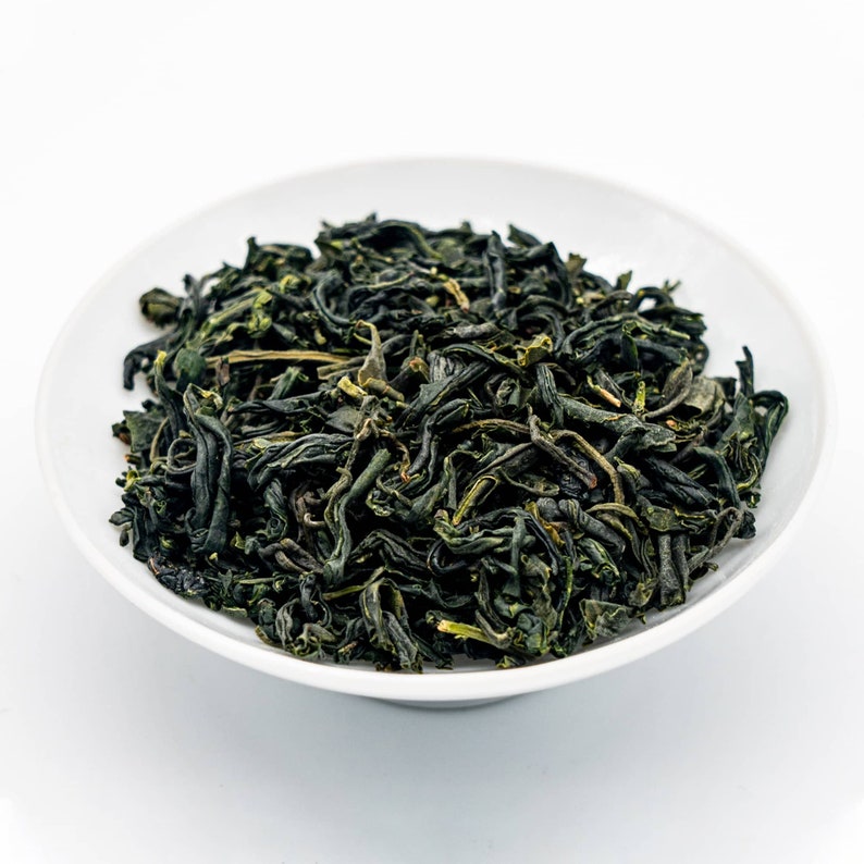 Puede incluir: Un cuenco de cer&aacute;mica blanco lleno de hojas de t&eacute; verde secas. Las hojas de t&eacute; son de color verde oscuro con algunos reflejos verde amarillentos m&aacute;s claros. El cuenco es redondo y est&aacute; sobre un fondo blanco.