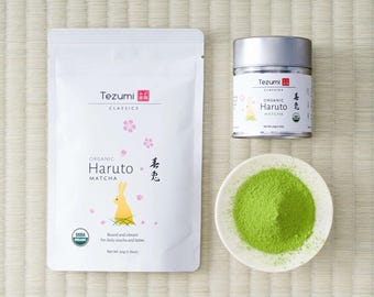 May include: A white pouch of Tezumi Classics Organic Haruto Matcha with a yellow rabbit illustration, a matching silver metal tin, and a small white bowl filled with vibrant green matcha powder. The packaging displays the text Organic Haruto Matcha.