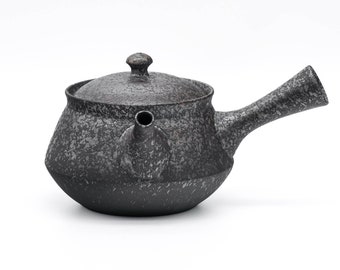 May include: A black ceramic teapot with a speckled finish and a rounded body. The teapot has a lid with a small knob and a spout on the side. The teapot has a long, curved handle.