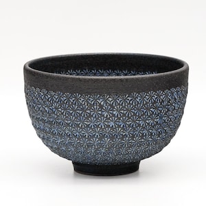 May include: A black ceramic bowl with a textured rim and a patterned exterior. The bowl's exterior features a repeating blue floral design. The bowl sits on a small, circular base. The background is white.