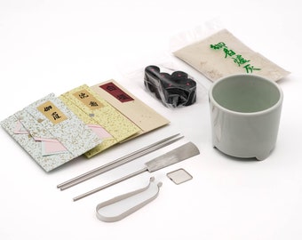 May include: A set of Japanese incense burning tools, including a white ceramic incense burner, a small bag of incense powder, a bag of black incense cones, three incense sticks, a metal incense stick holder, a metal incense stick cutter, and three small packets of incense with Japanese characters.