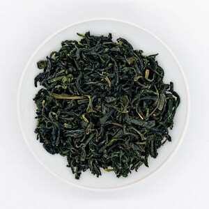 Puede incluir: Vista superior de hojas de t&eacute; sueltas en un cuenco de cer&aacute;mica blanco. Las hojas de t&eacute; son de color verde oscuro, con tonos verdes m&aacute;s claros. Las hojas est&aacute;n fuertemente enrolladas, lo que sugiere alta calidad.