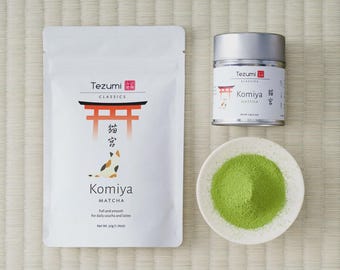 May include: A white pouch and a silver metal tin of Tezumi Komiya matcha, both featuring a red torii gate and a calico cat illustration. A small white bowl contains a mound of vibrant green matcha powder, net weight 50g (1.76oz).