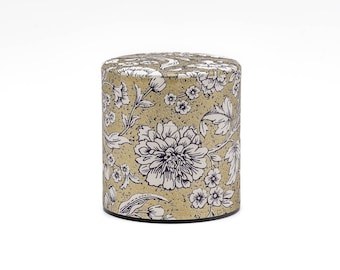 May include: A cylindrical gold tea tin with a floral pattern in white and purple. The tin has a black base.