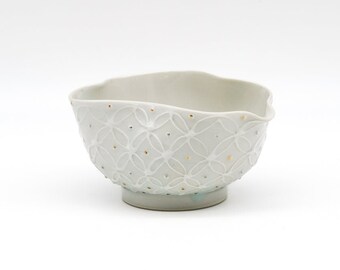 May include: A white ceramic bowl with a delicate, embossed pattern and gold accents. The bowl has a slightly irregular shape and a rounded base.