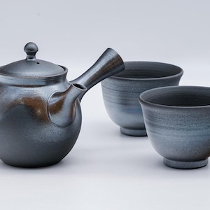 May include: A dark gray ceramic teapot with a lid and a spout, along with two matching gray ceramic teacups. The teapot and cups have a subtle blue glaze.