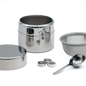Könnte beinhalten: Ein Set aus Edelstahl-Teezubehör, bestehend aus einer runden Teedose mit Deckel, einem Sieb und einem kleinen Löffel.