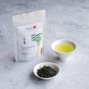 Puede incluir: Una bolsa blanca de t&eacute; Kamairicha "Tezumi Classics" con ilustraci&oacute;n de bamb&uacute;, un cuenco blanco peque&ntilde;o con t&eacute; suelto y una taza blanca llena de t&eacute; verde. El texto de la bolsa dice "Takamura" y "Round and silky pan-fired green tea."